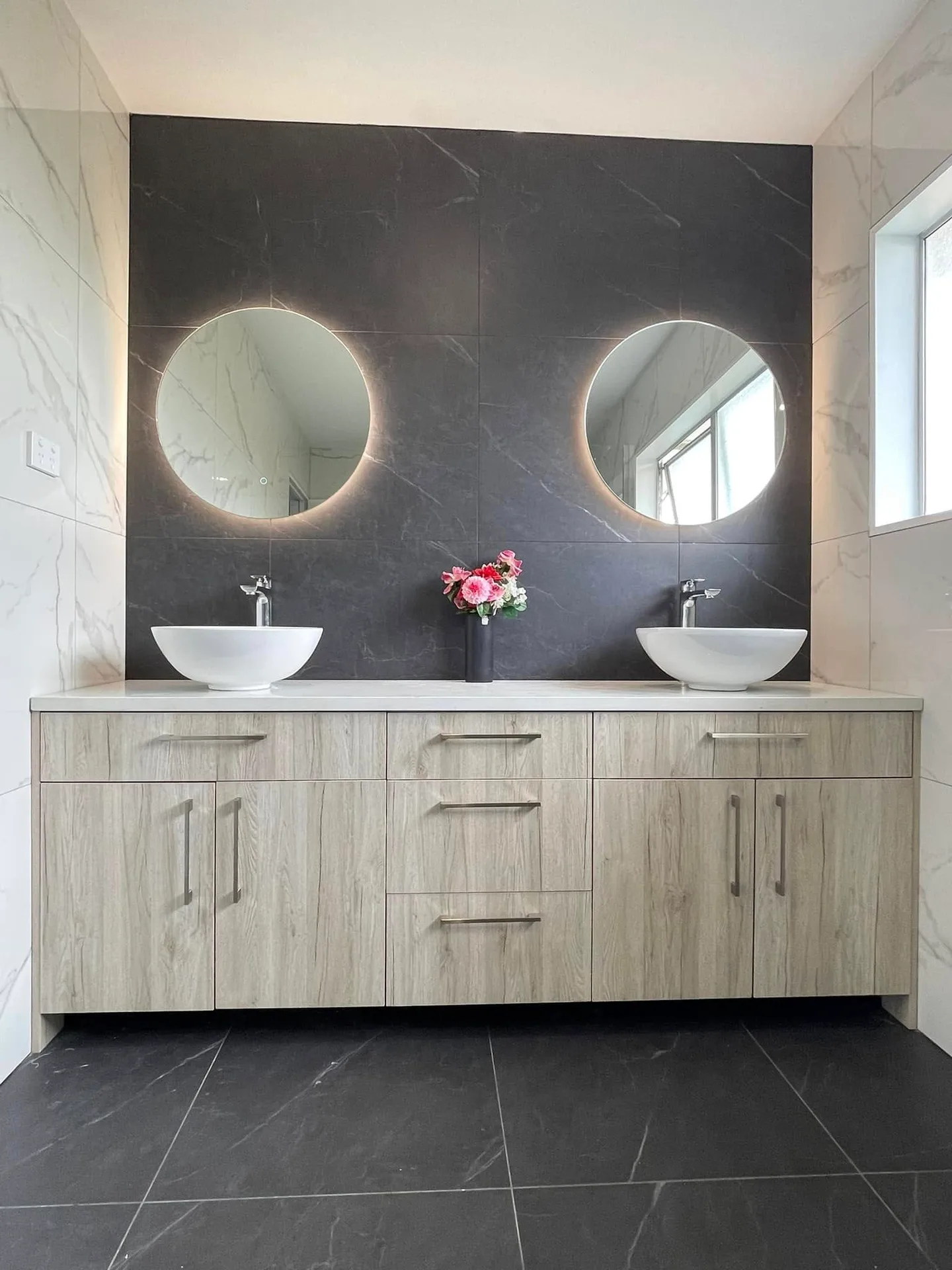 Double basin bathroom with round backlit mirrors and dark marble feature wall